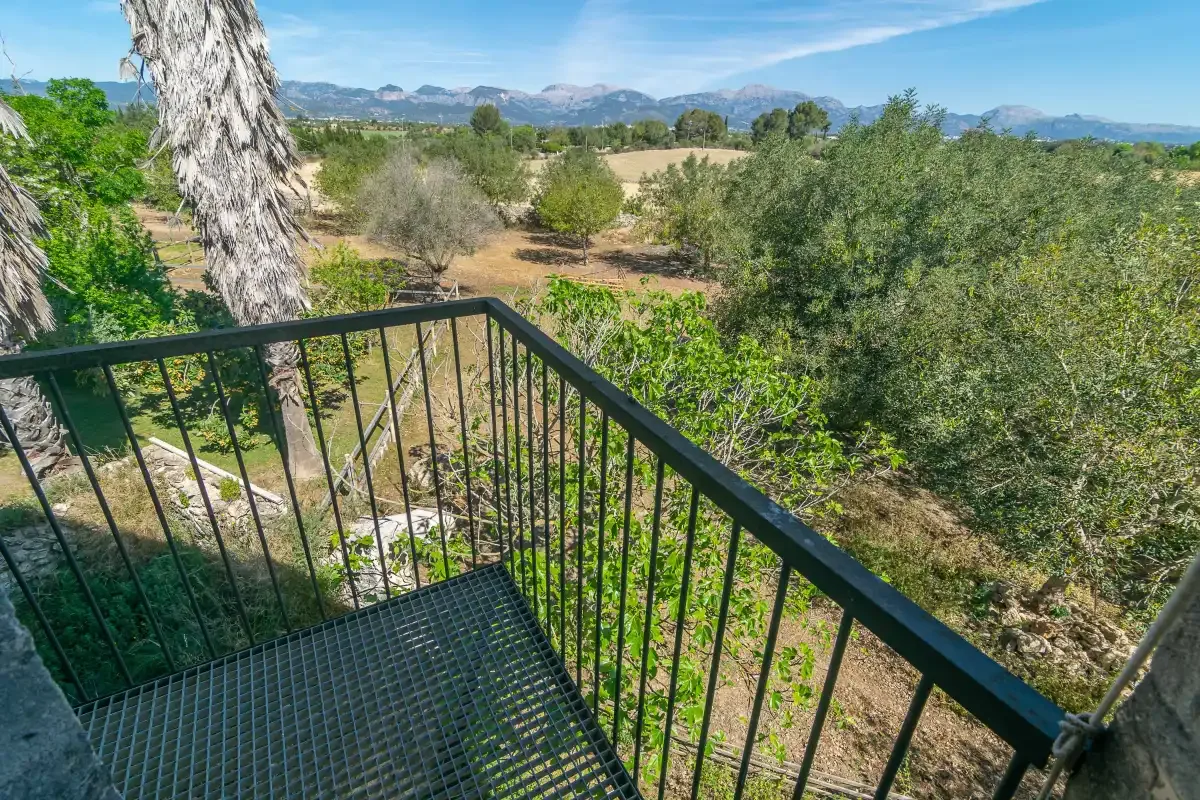 Un balcón metálico da a árboles verdes y montañas lejanas.