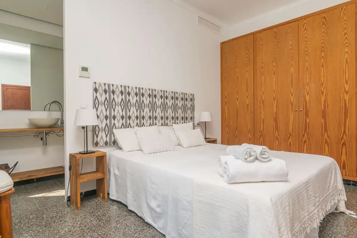 Un dormitorio luminoso con una cama blanca, un cabecero estampado y un armario de madera.