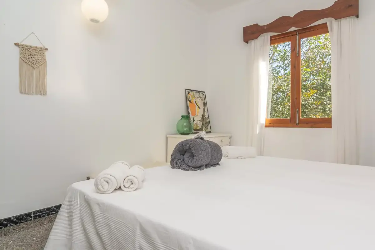 Un dormitorio luminoso con ropa de cama blanca y una ventana con vista a árboles frondosos.