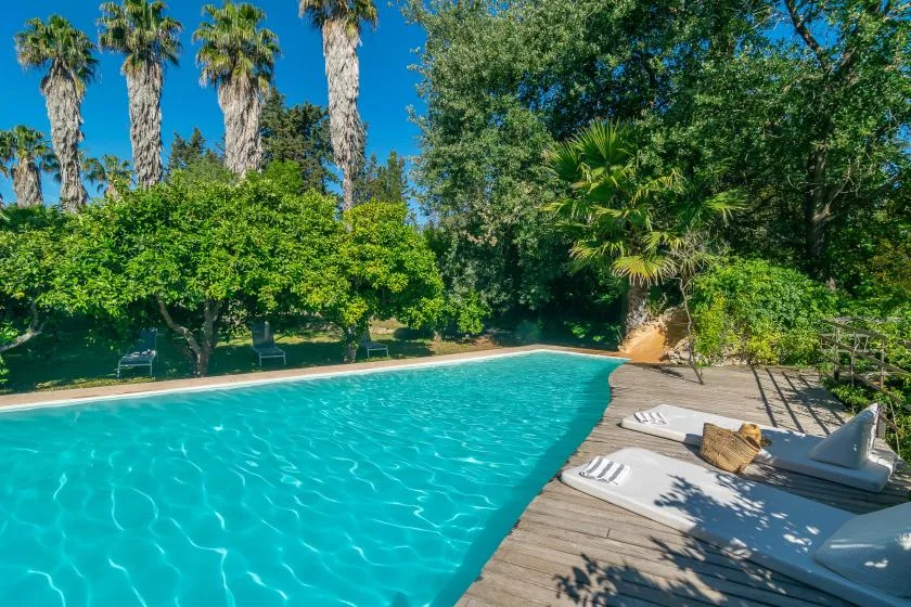 Una piscina turquesa está junto a una terraza de madera con tumbonas y vegetación alrededor.
