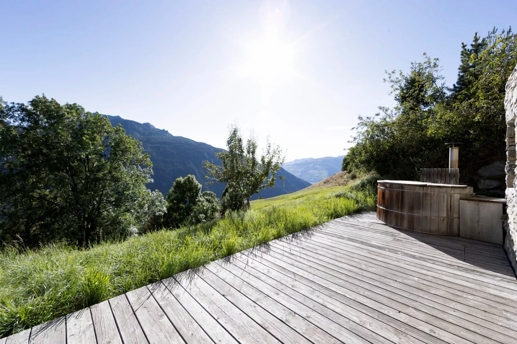 Una terraza de madera da a un valle montañoso verde, con una bañera exterior al borde.