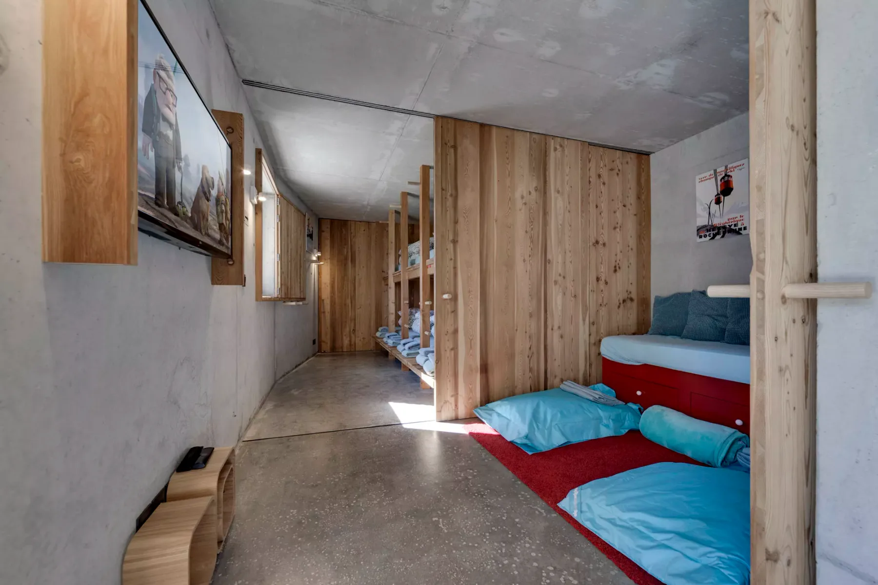 Una habitación estrecha con divisiones de madera, ropa de cama azul y piso de concreto.