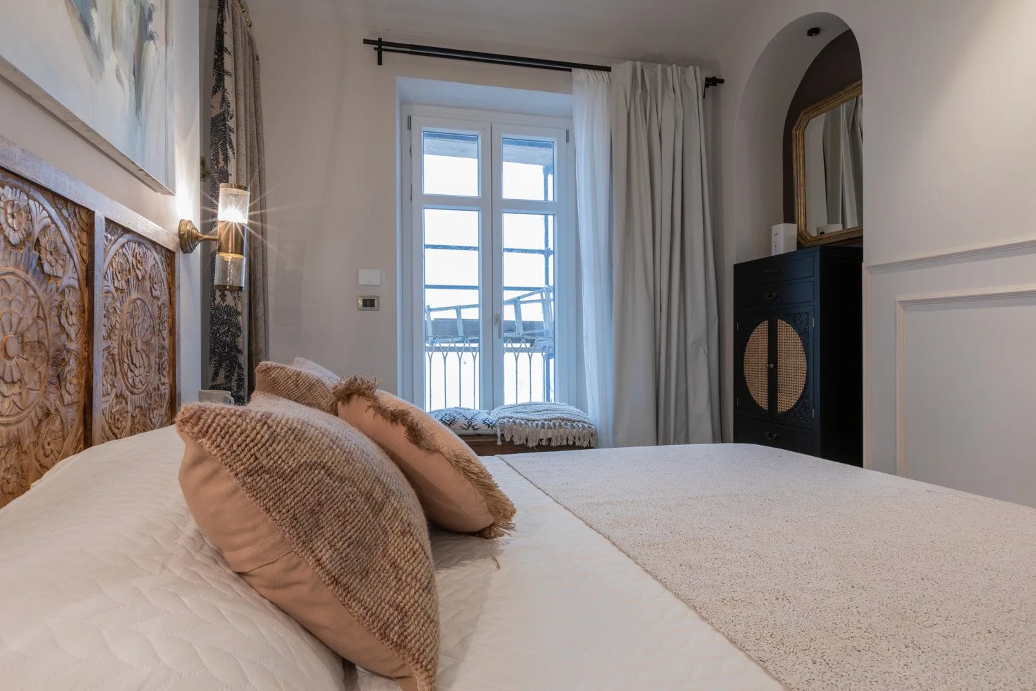 A bedroom with a bed, decorative pillows, and a tall window with curtains.