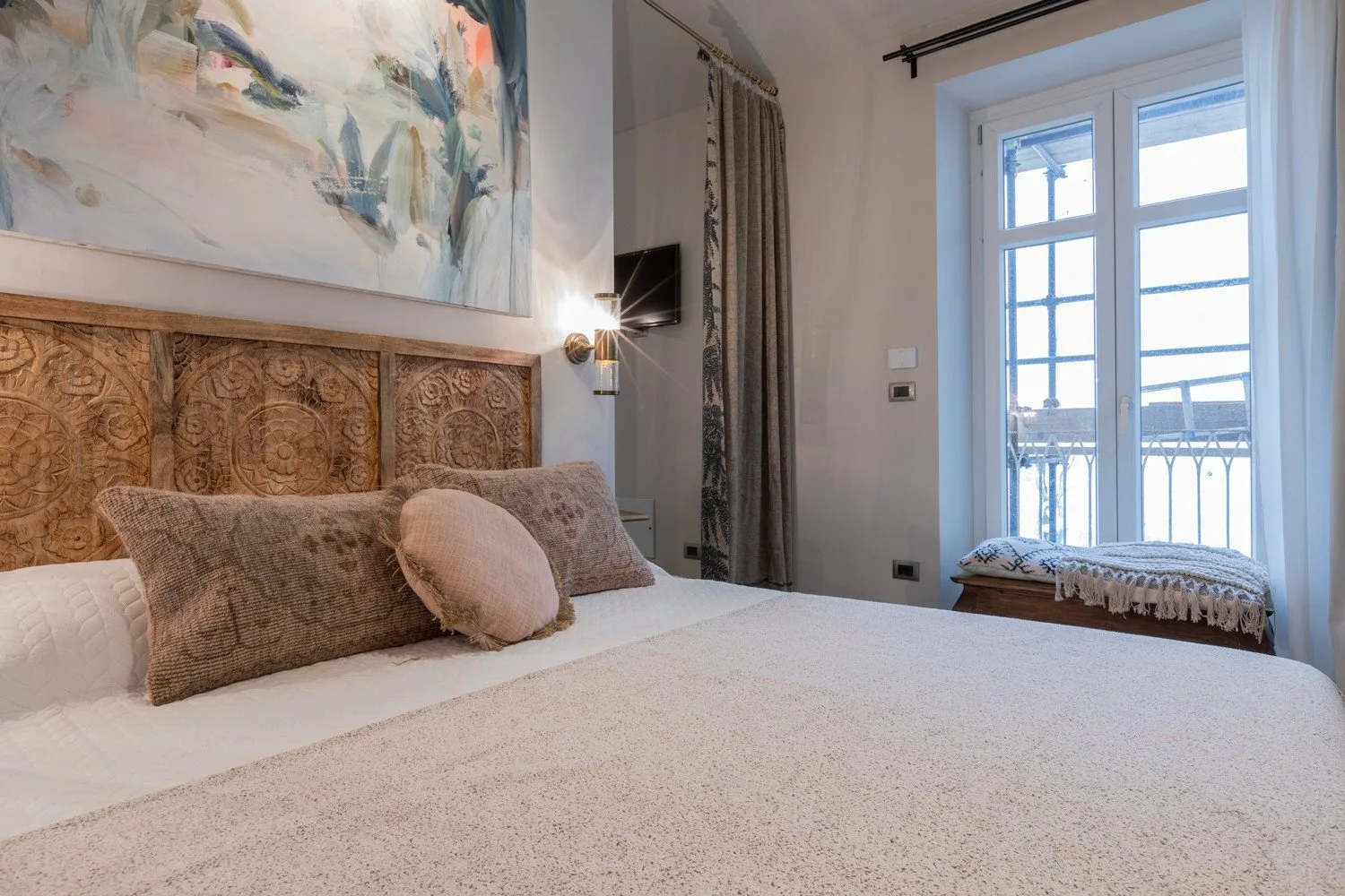 A bedroom with a bed beneath a large painting and beside a bright window.