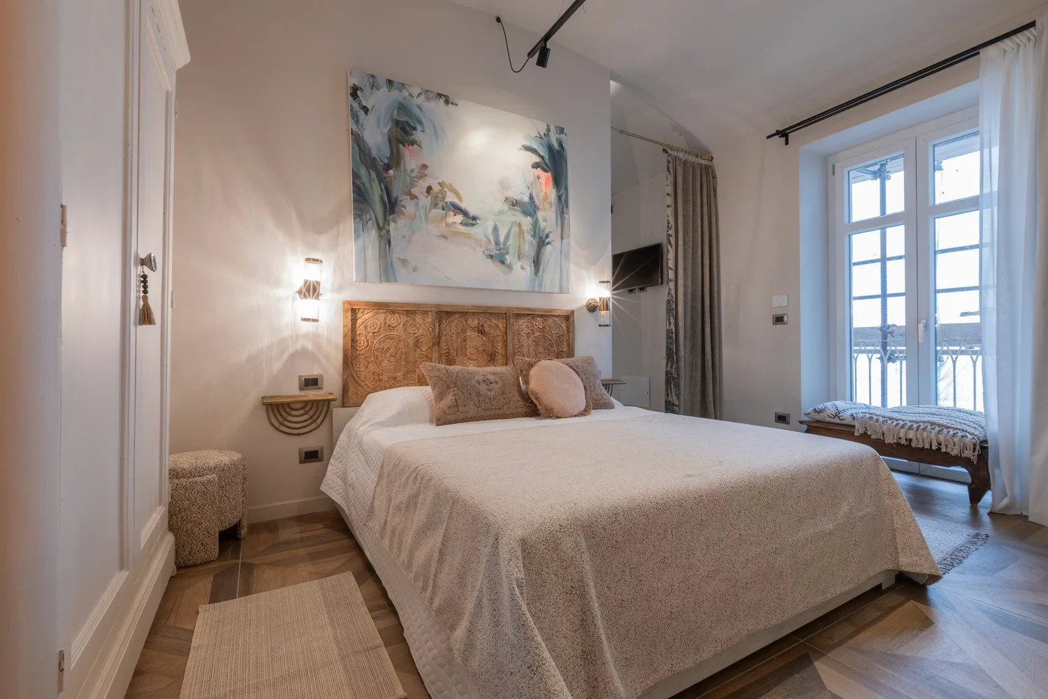 A bedroom with a bed beneath a colorful painting and tall windows.