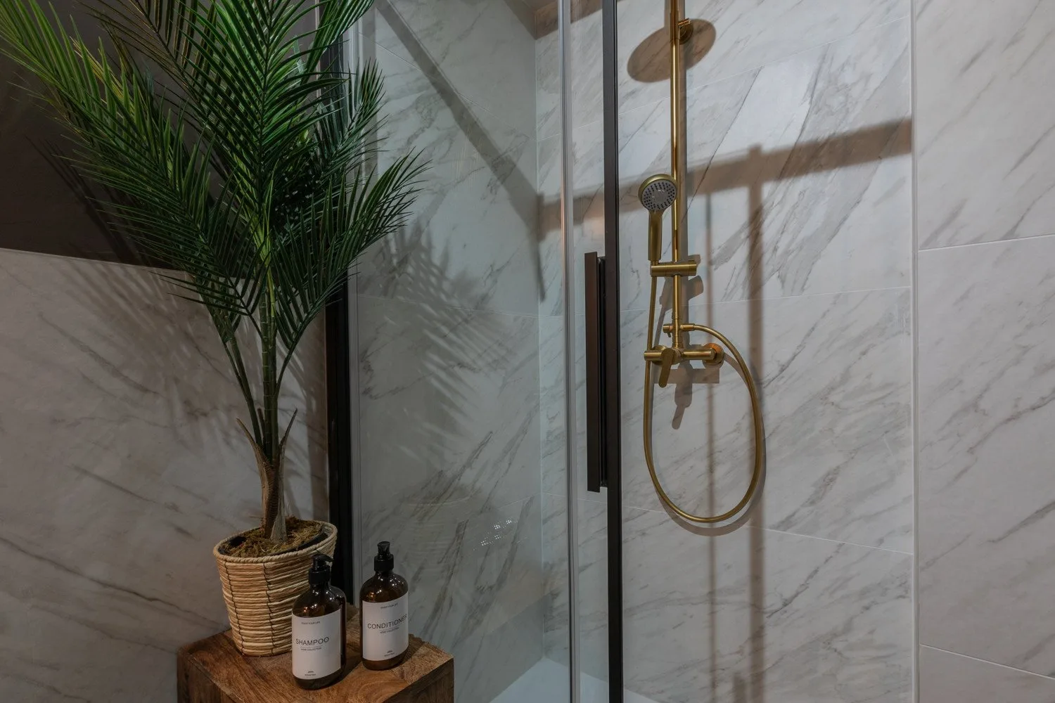A tiled bathroom shower with gold-colored fixtures and a potted plant nearby.