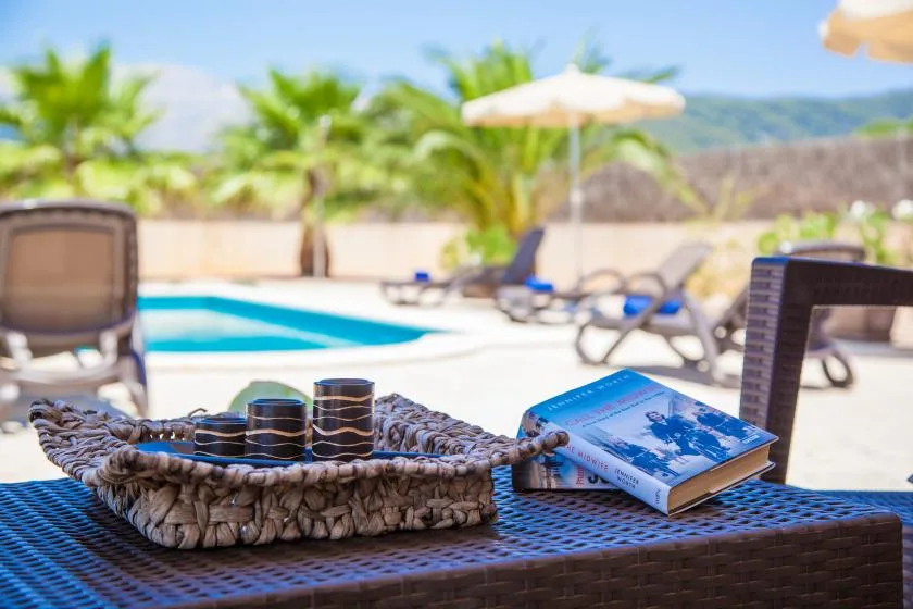 A poolside table holds a woven tray and book beside a swimming pool.