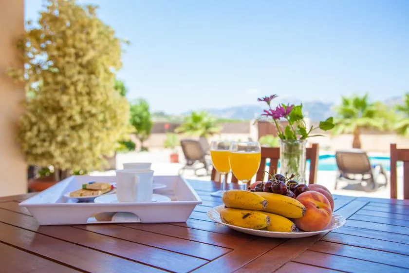 A sunny outdoor dining table holds fruit and orange drinks beside a pool.
