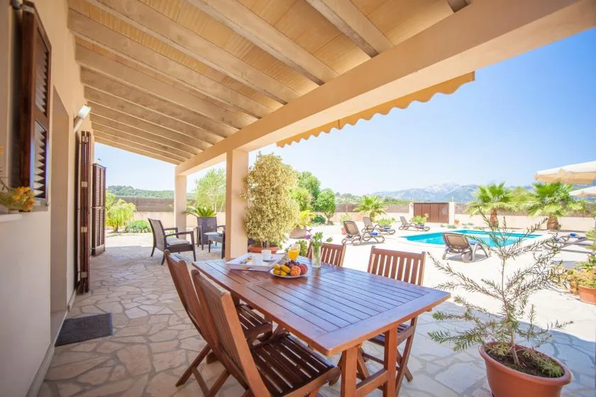 A covered outdoor dining area overlooks a swimming pool and sunny landscaped grounds.