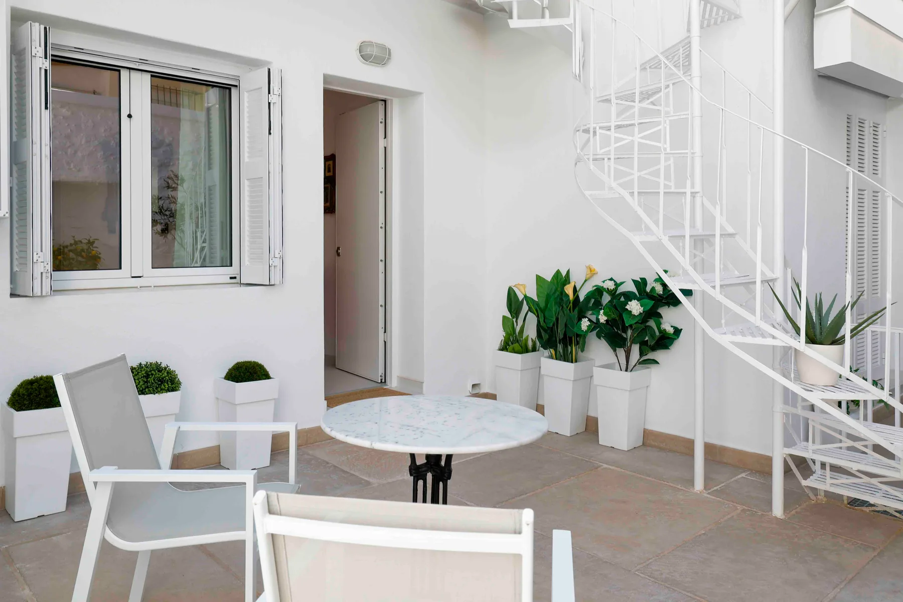 Un patio luminoso con asientos blancos, plantas en macetas y una escalera de caracol.
