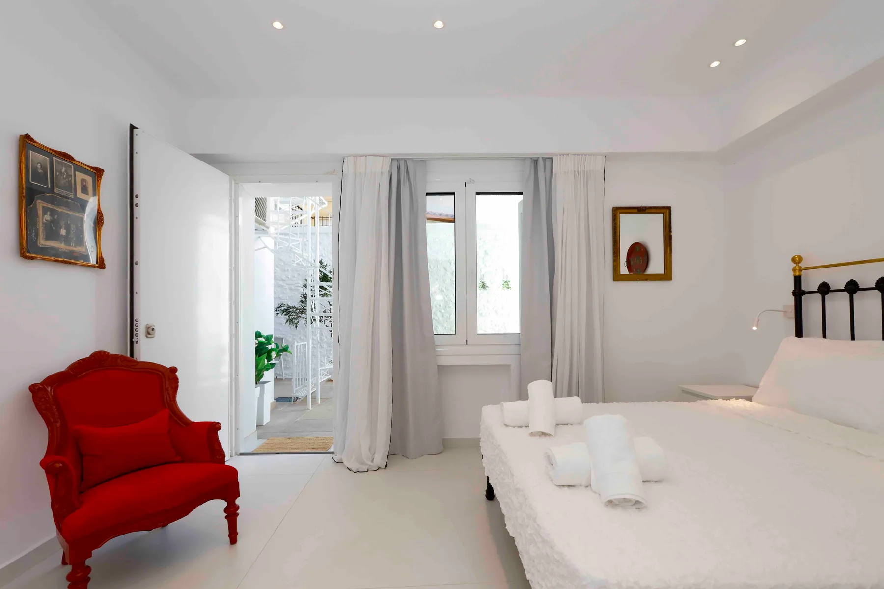 Un dormitorio luminoso con una cama blanca, cortinas grises y un sillón rojo.