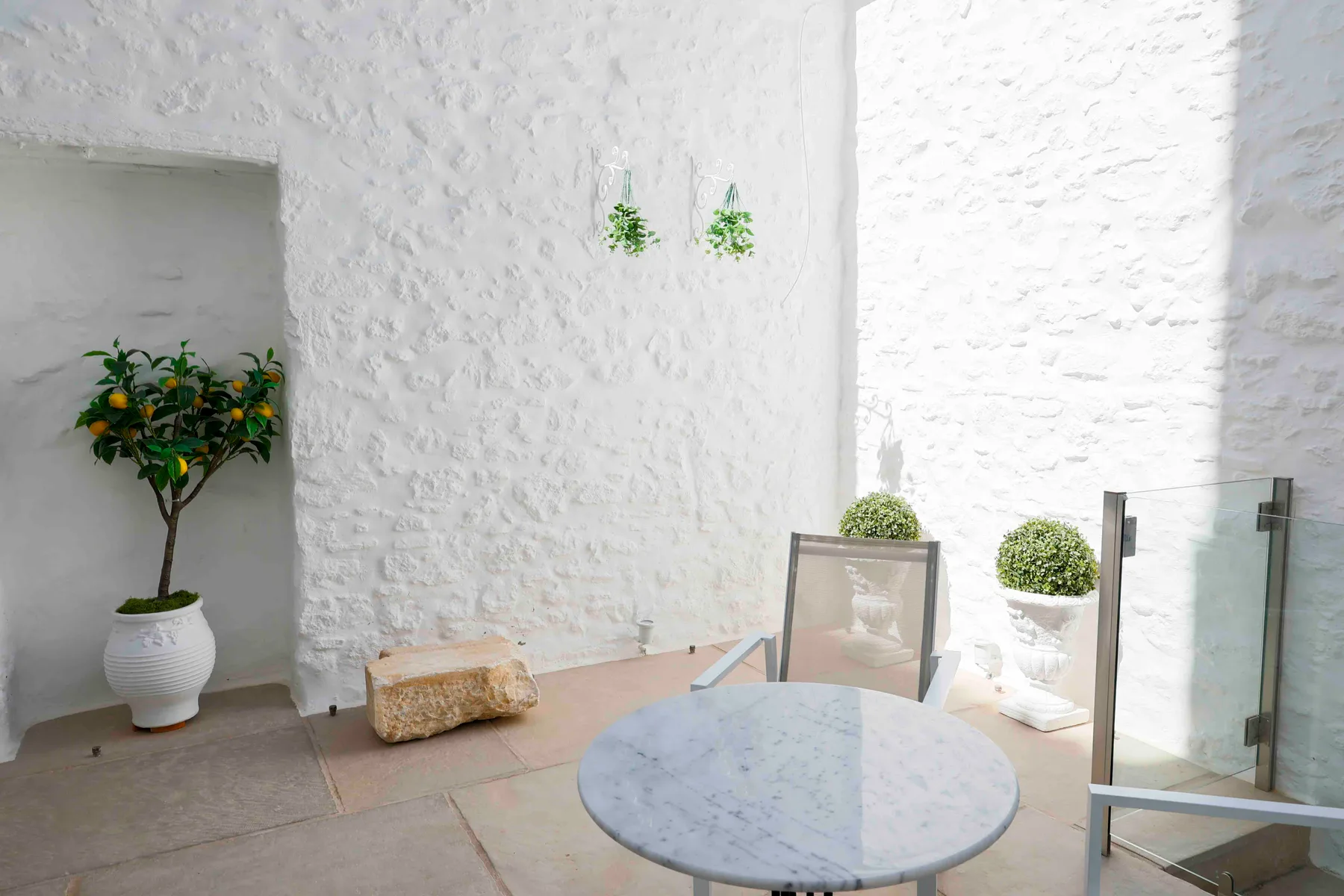 Una terraza luminosa de piedra blanca con una mesa redonda y plantas en macetas.