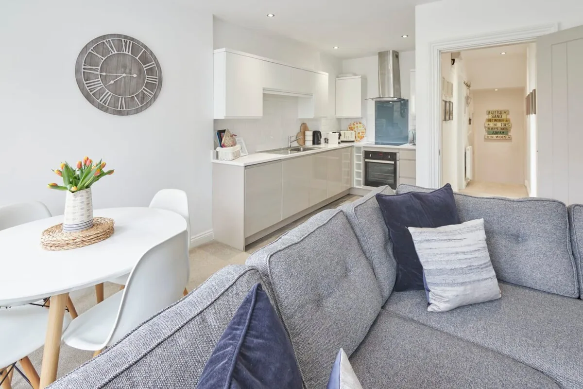 A bright open-plan living area with a gray sofa, dining table, and white kitchen.