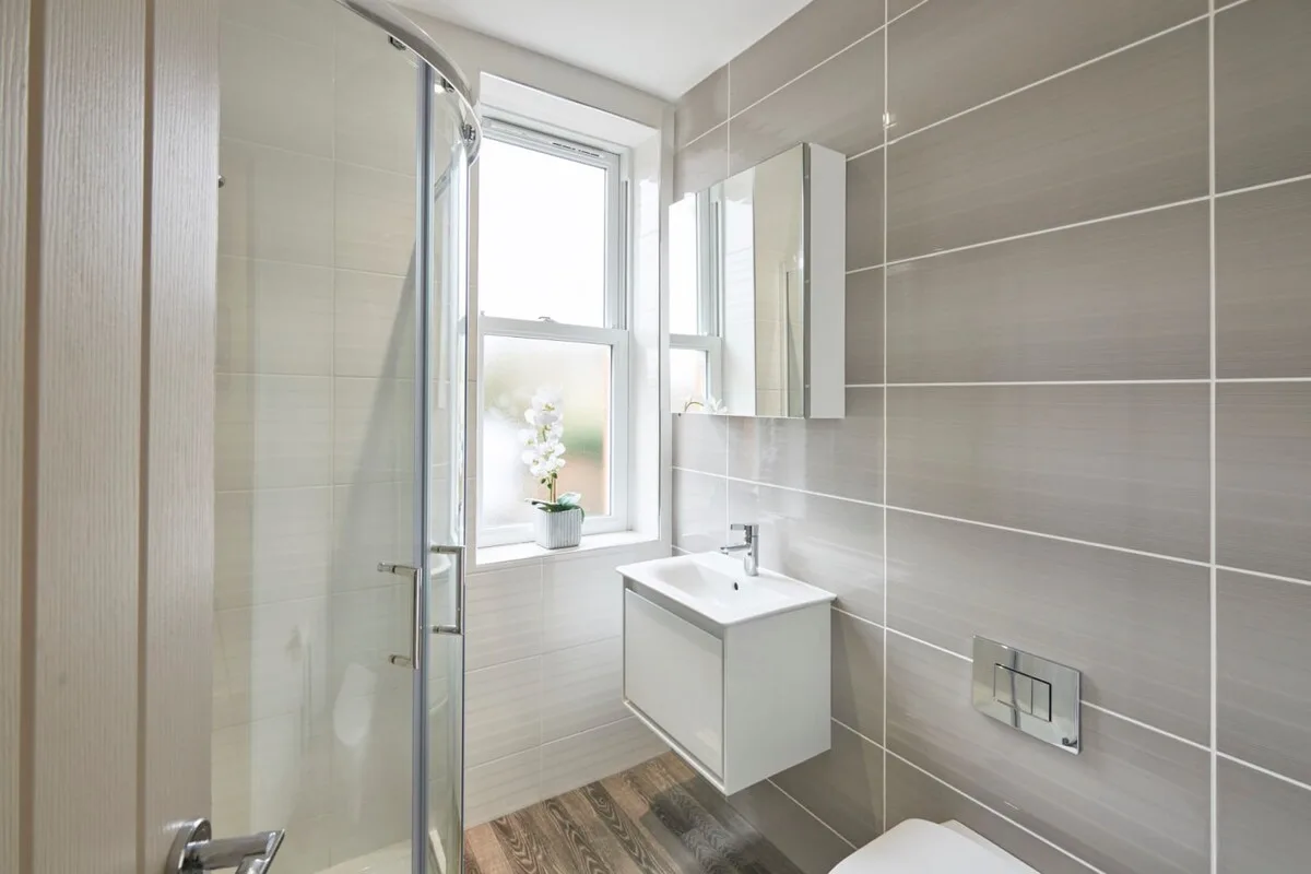 A bright tiled bathroom with a glass shower, window, and wall-mounted sink.