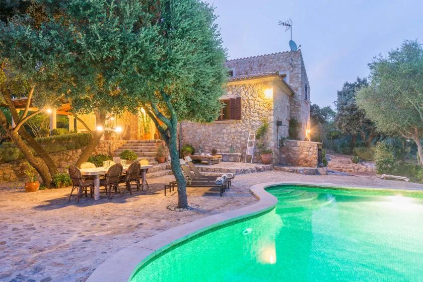 A stone house and outdoor dining area beside a brightly lit swimming pool.