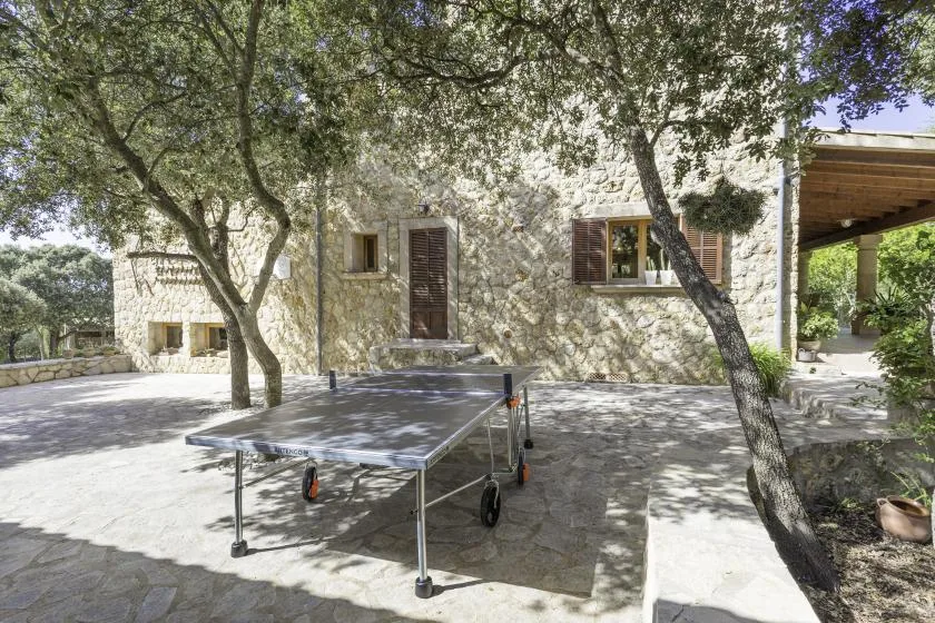 A stone building overlooks a paved patio shaded by trees, with a table tennis table.