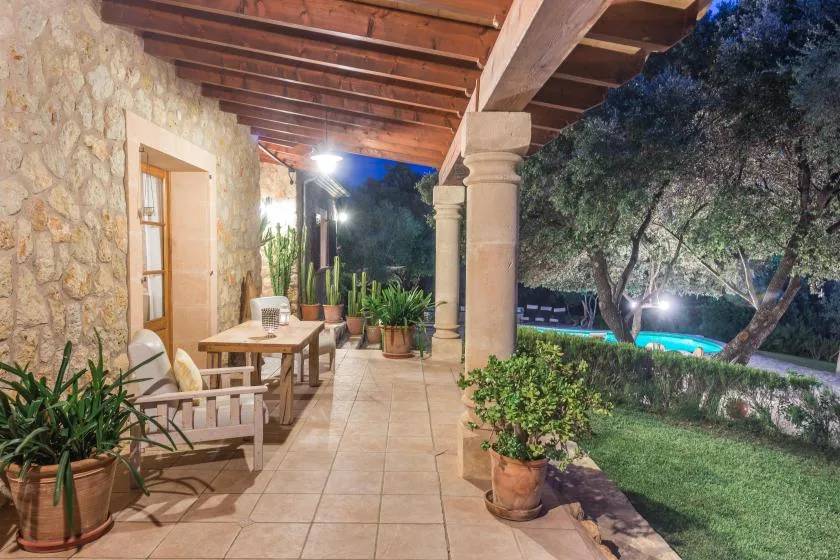 A covered stone patio with outdoor seating and potted plants beside a garden.