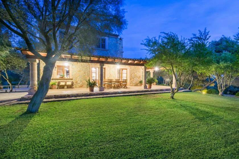 A warmly lit stone house stands beside a broad lawn and mature trees at dusk.