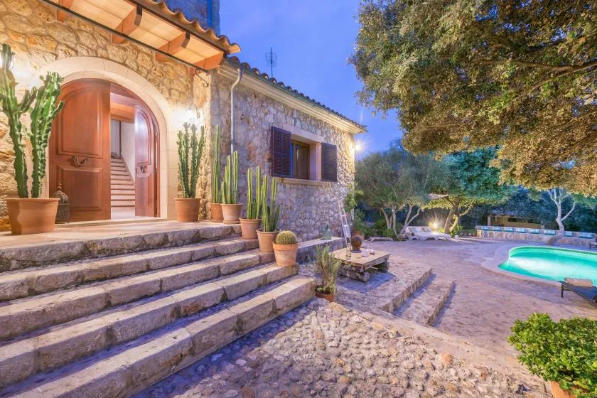 A stone house opens onto a paved courtyard with a pool and surrounding trees.