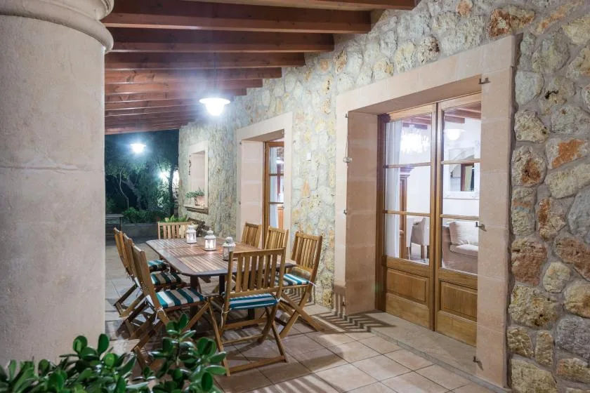 A covered stone terrace has a wooden dining table and chairs beside glazed doors.