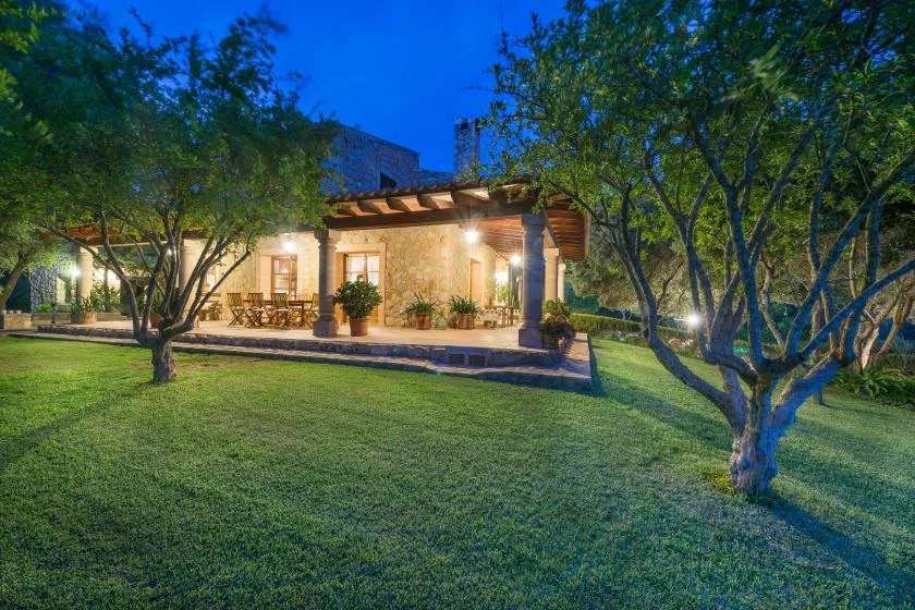A warmly lit house with a covered patio sits beside a broad green lawn.