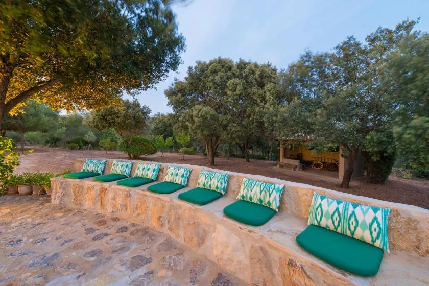 An outdoor stone seating area with green cushions sits beneath leafy trees.