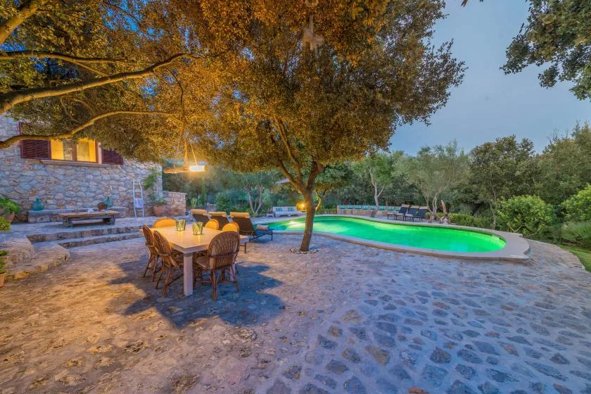 A stone patio with outdoor dining seating overlooks a swimming pool beneath trees.