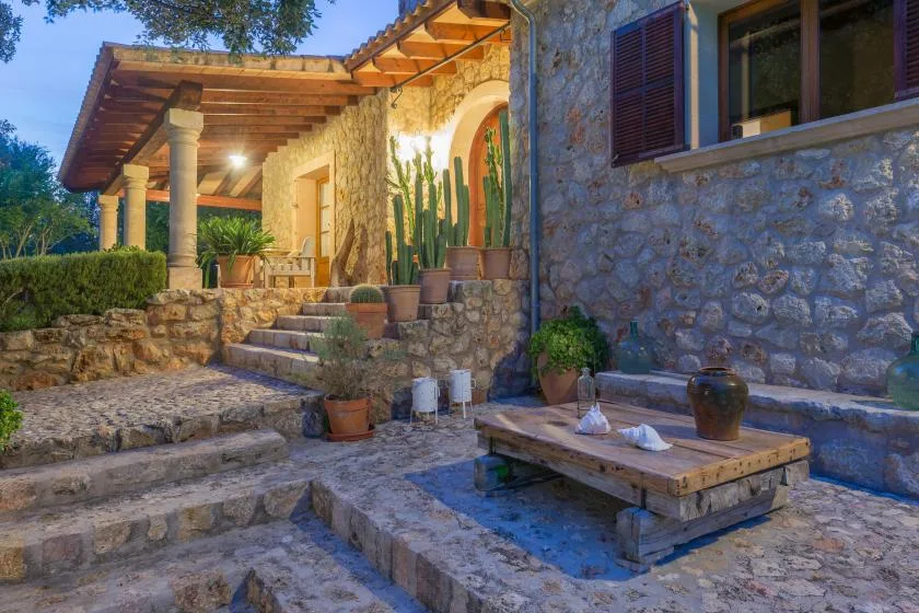 A stone patio with a low wooden table and potted plants beside a house.
