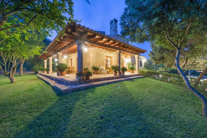 A warmly lit house with a covered patio sits amid a green lawn and trees.