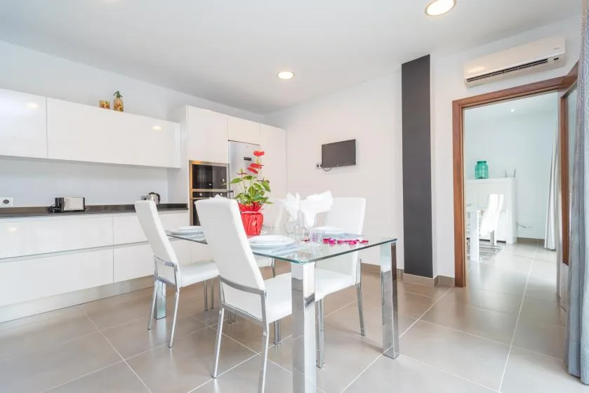 Un comedor luminoso con mesa de vidrio y asientos blancos junto a una cocina moderna.