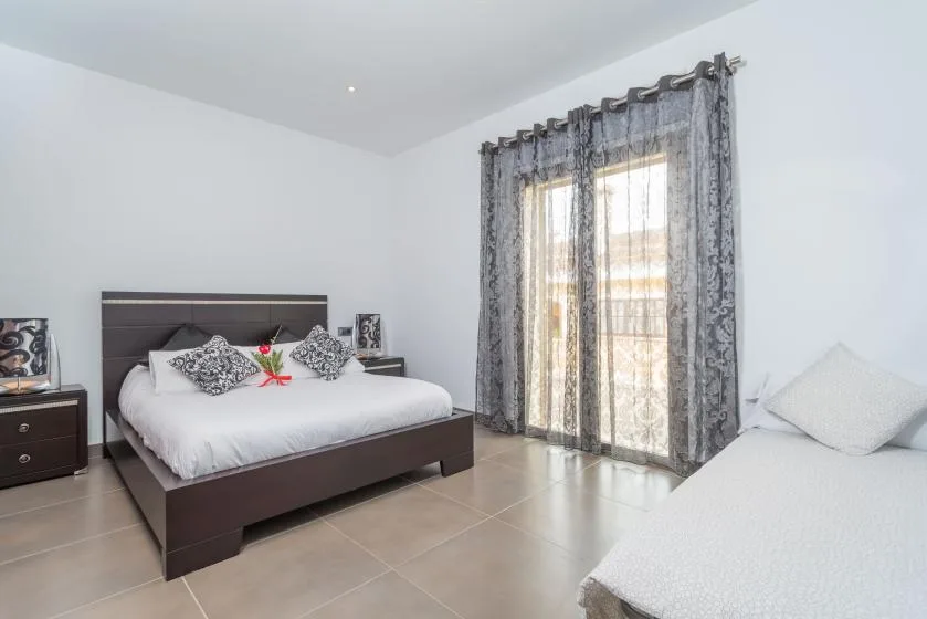 Un dormitorio luminoso con una cama oscura y cortinas transparentes junto a la ventana.