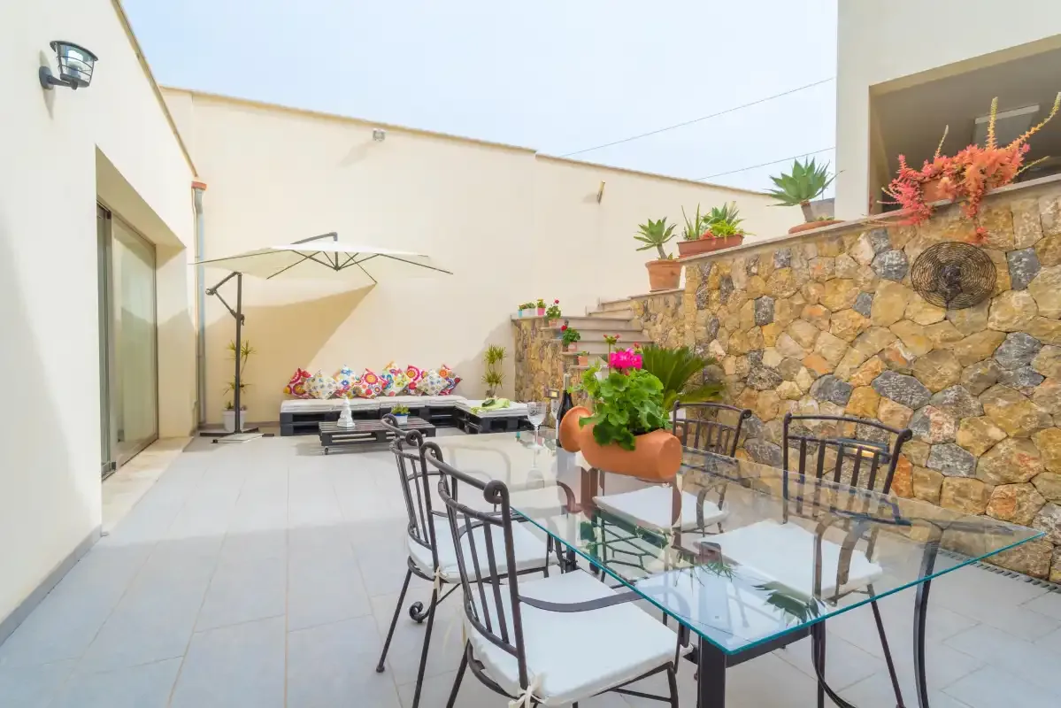 Un patio al aire libre con una mesa de vidrio, asientos metálicos y plantas en macetas.