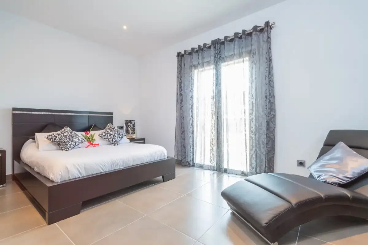 Un dormitorio con una cama y una chaise longue junto a puertas de vidrio con cortinas.