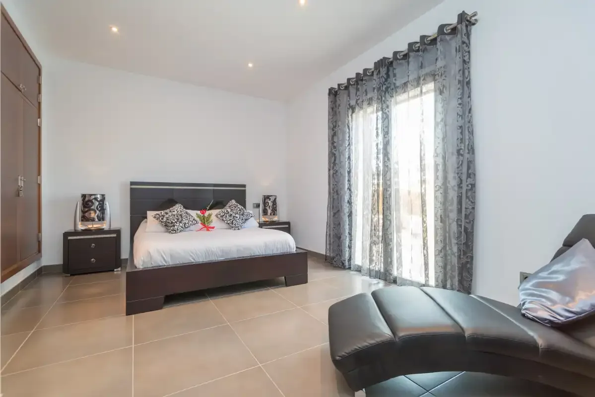 Un dormitorio luminoso tiene una cama junto a ventanas altas con cortinas.
