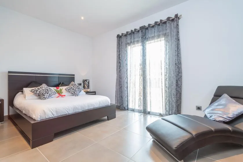 Un dormitorio con una cama y una chaise longue junto a ventanas altas con cortinas.