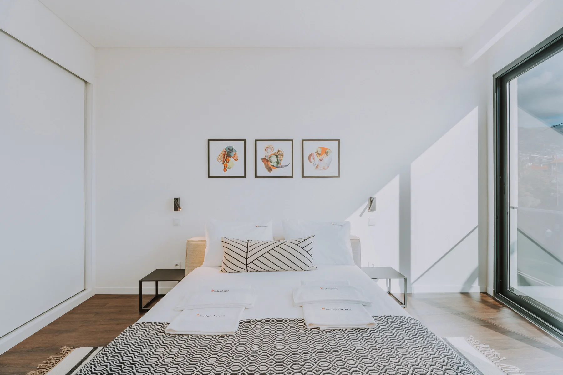 Dormitorio luminoso con una cama tendida y cuadros enmarcados sobre ella.