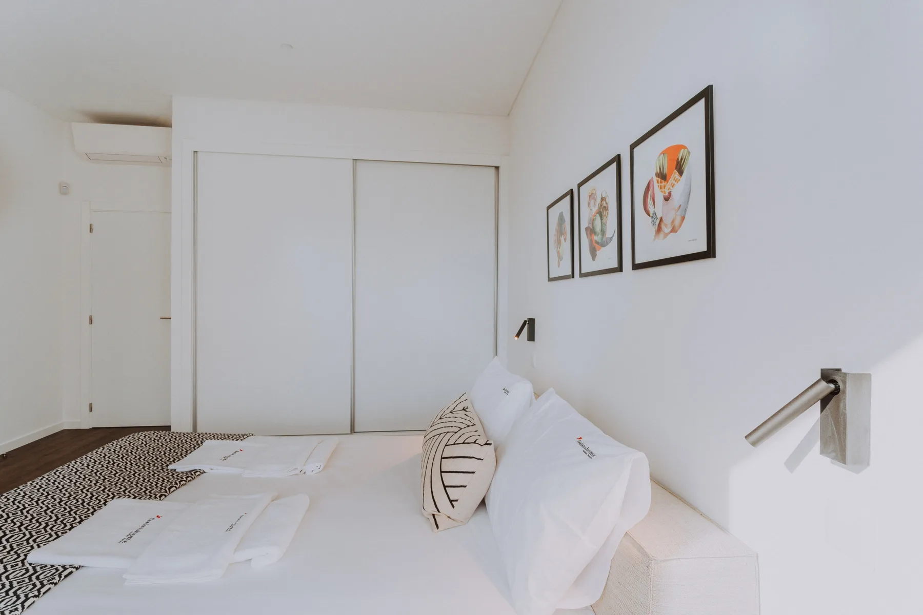 Un dormitorio luminoso con ropa de cama blanca, cuadros enmarcados y puertas correderas de armario.