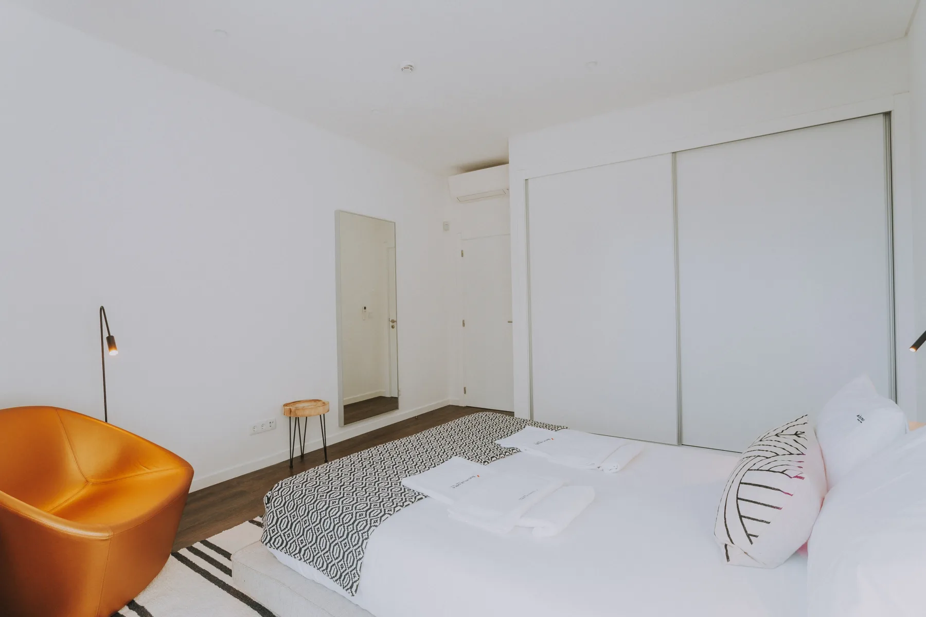 Un dormitorio luminoso con una cama blanca, ropa de cama estampada y un sillón naranja.