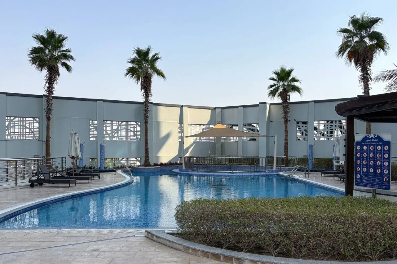 Una piscina al aire libre junto a una terraza de baldosas y palmeras.