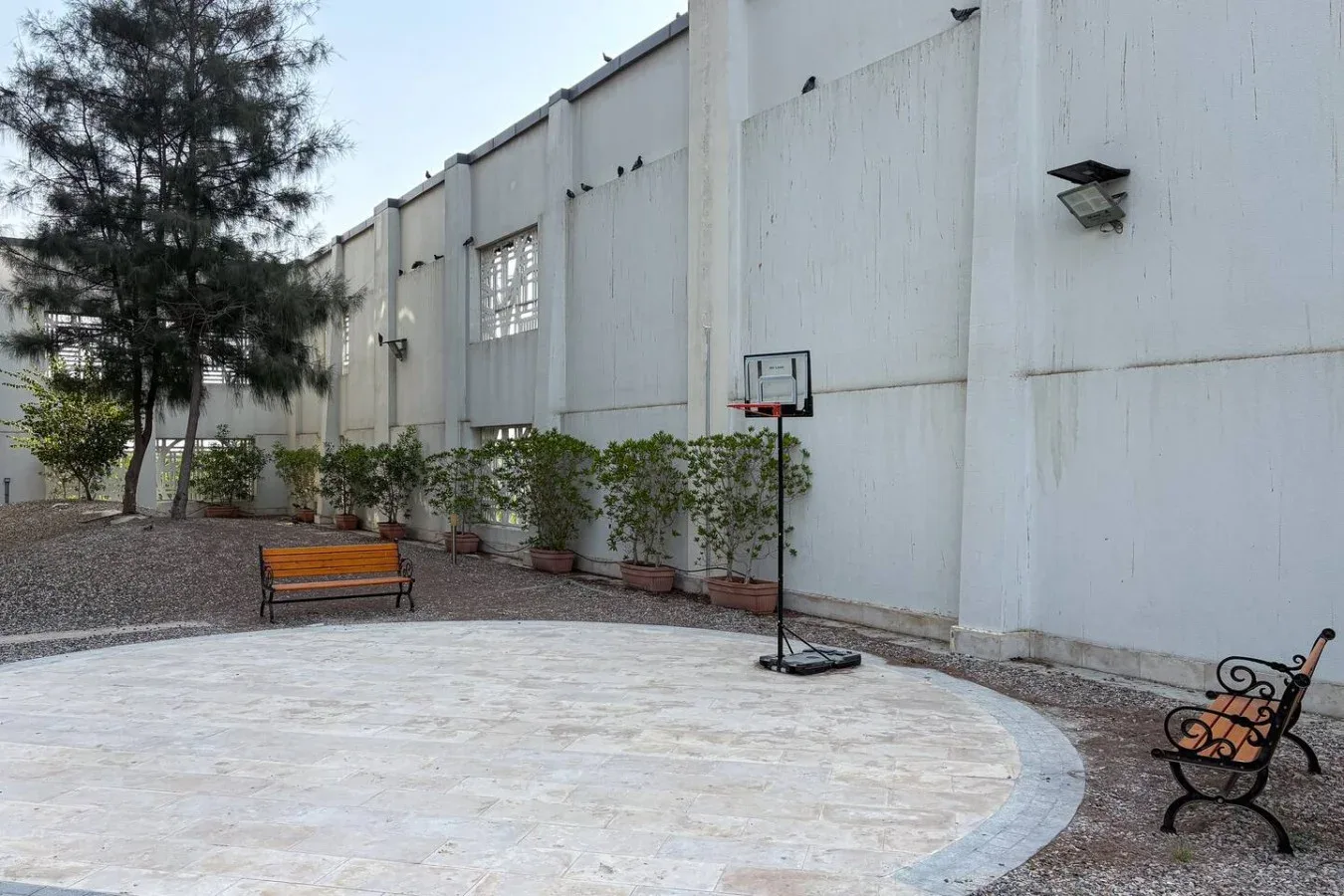 Una cancha de baloncesto al aire libre junto a un edificio blanco alto, con una canasta y bancos.