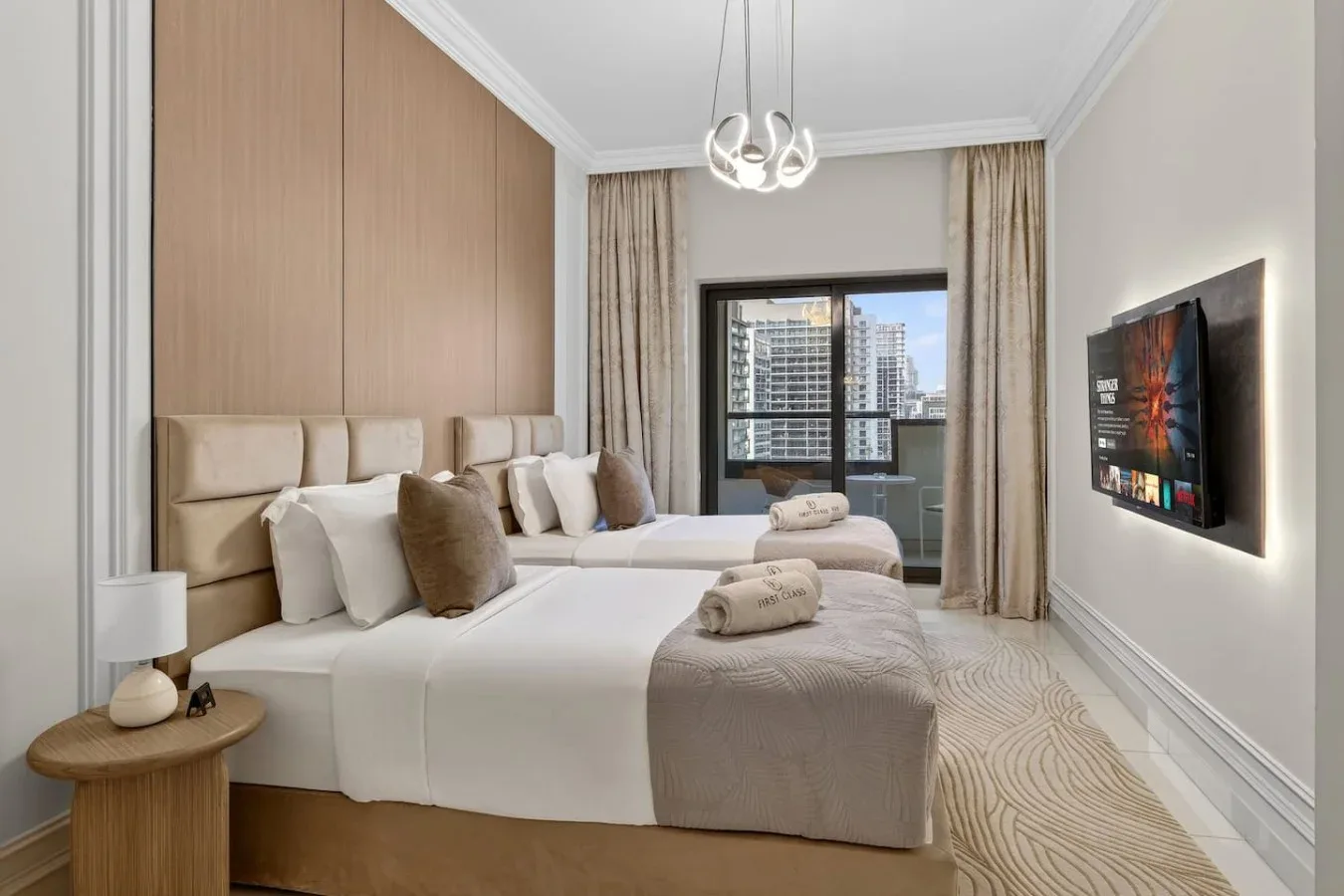 Un dormitorio luminoso con dos camas, ropa de cama neutra y una ventana con edificios cercanos a la vista.