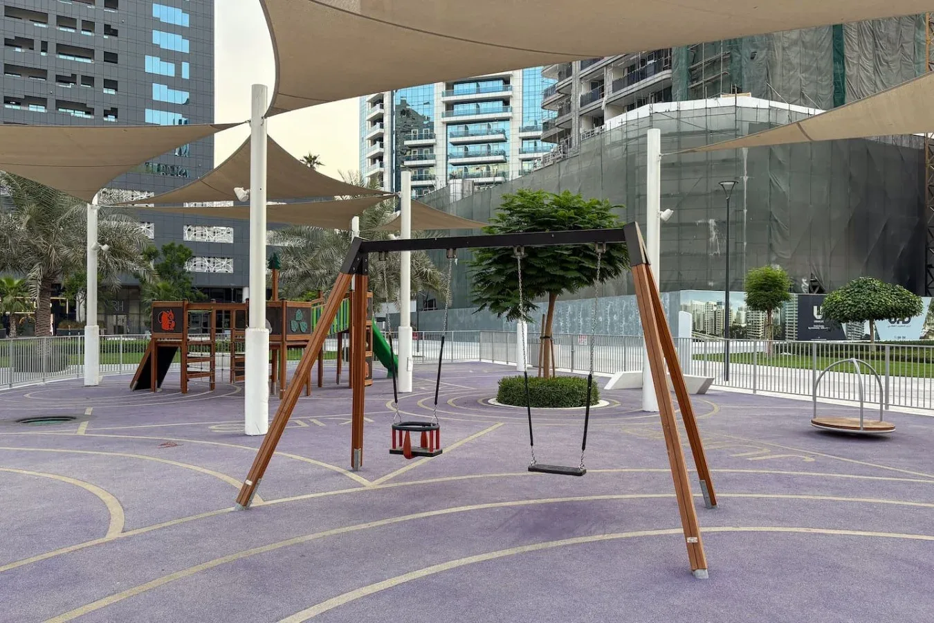 Un parque infantil al aire libre con columpios bajo toldos entre edificios modernos.