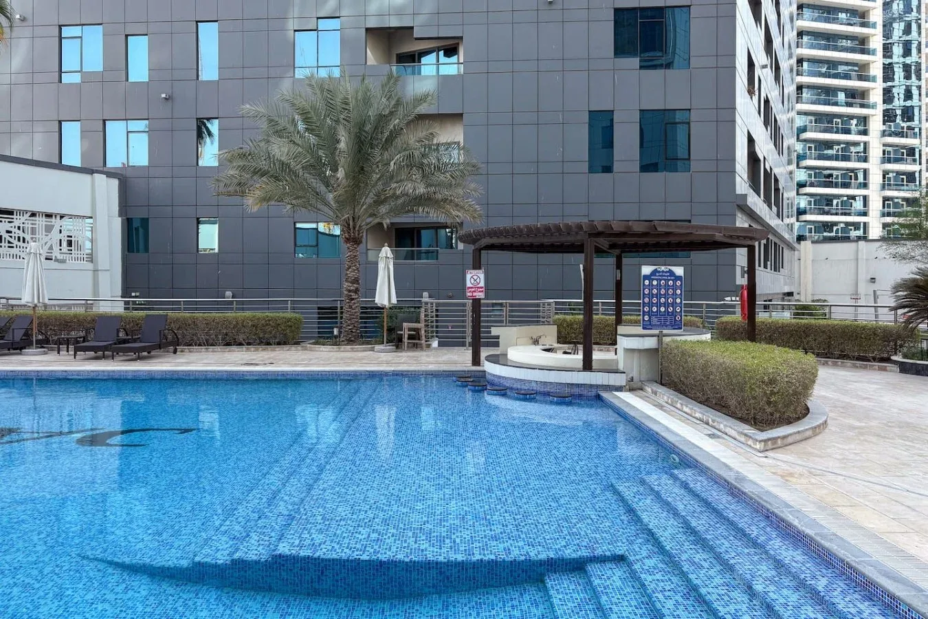 Una piscina al aire libre junto a una terraza pavimentada y edificios.