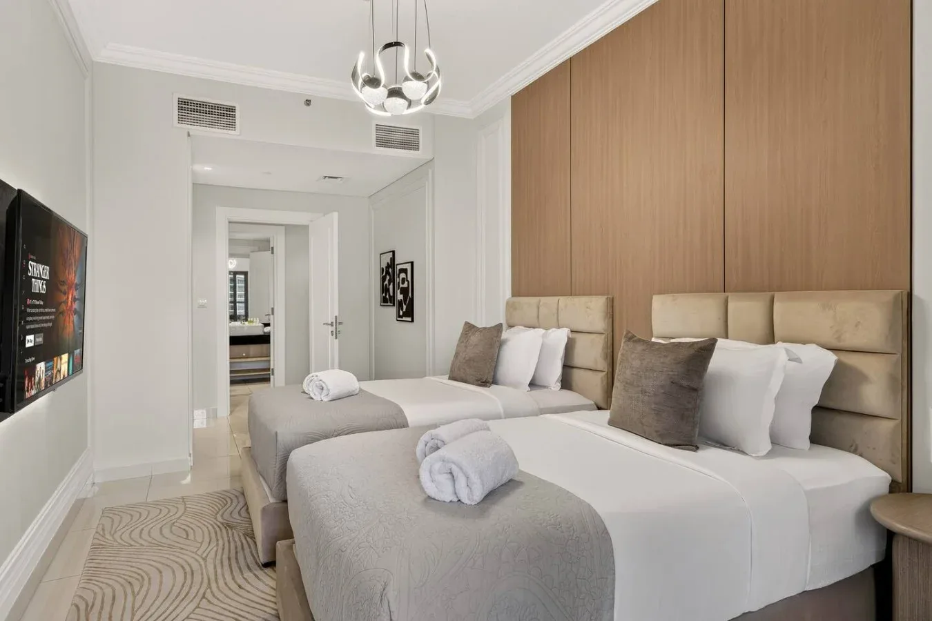 Un dormitorio luminoso con camas, ropa de cama neutra y una pared revestida de madera.