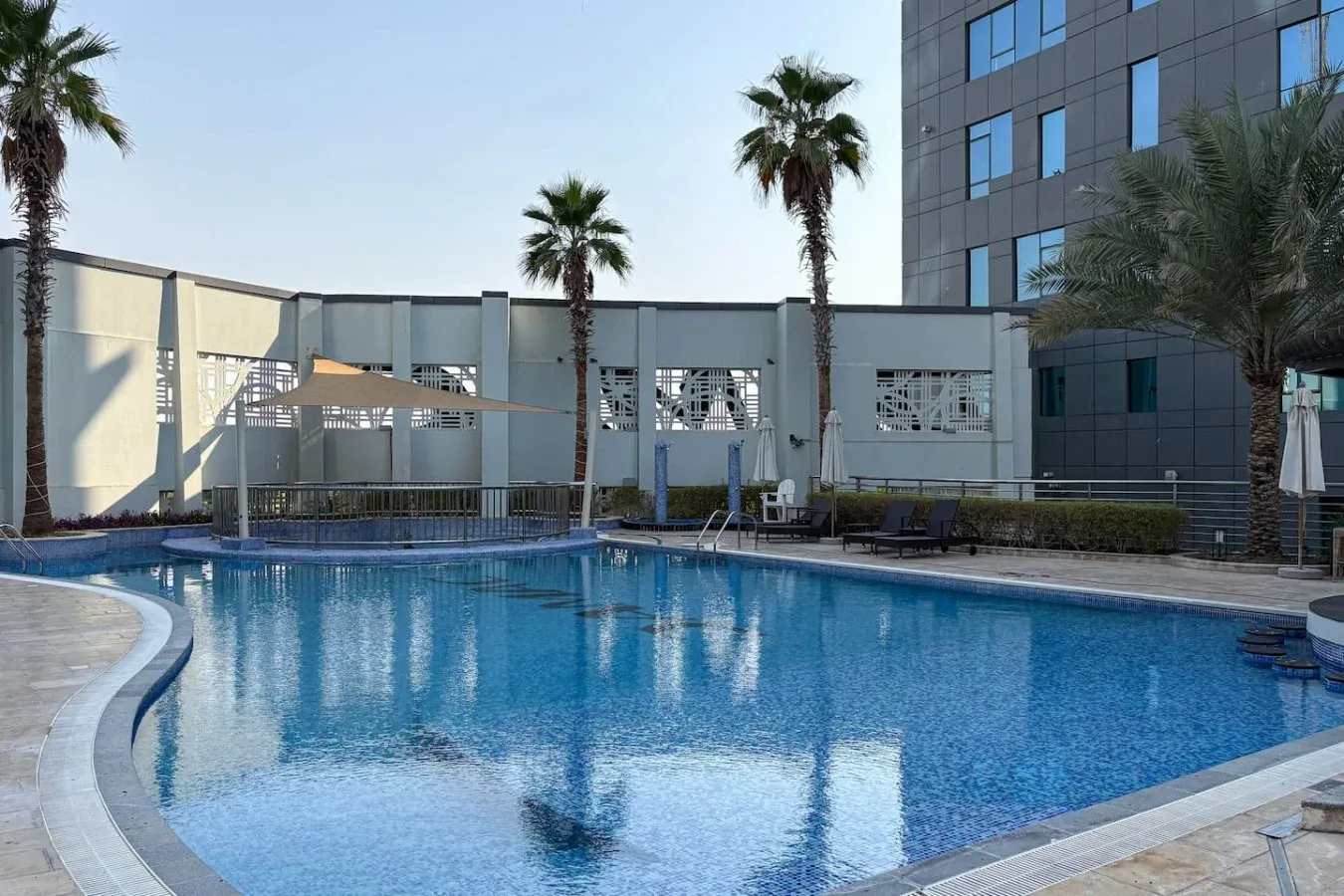 Una piscina al aire libre junto a un edificio moderno, con palmeras en el borde.