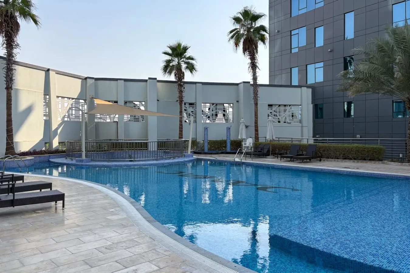 Una piscina al aire libre junto a un edificio moderno, con palmeras junto a la zona de piscina.
