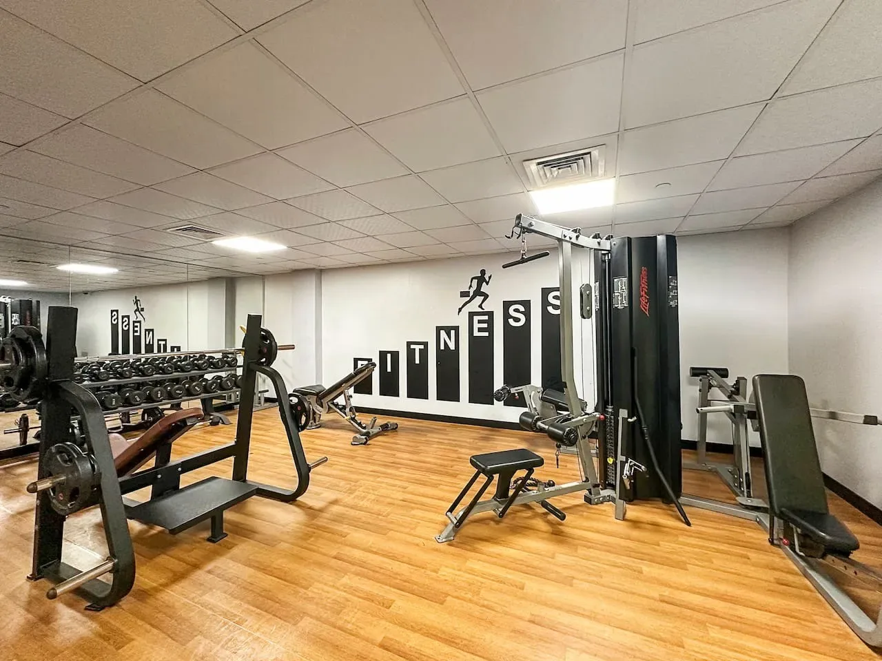Un gimnasio con máquinas de pesas y bancos distribuidos en una sala con suelo de madera.