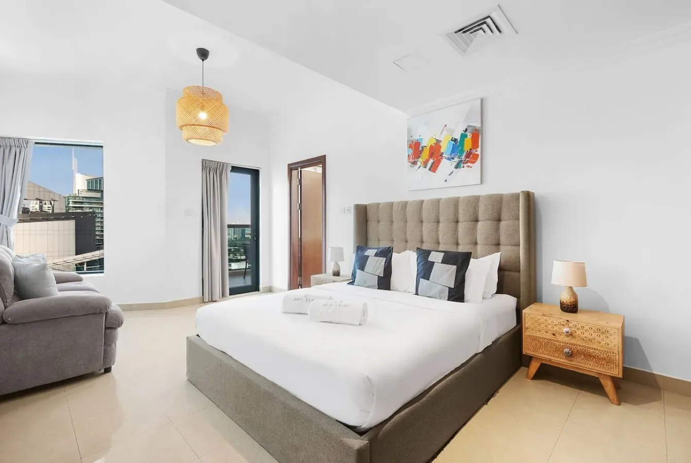 Un dormitorio luminoso con una cama grande, cabecero acolchado y una zona de asientos junto a la ventana.