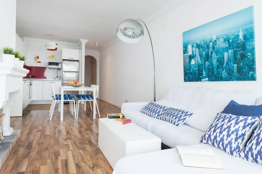 Una sala luminosa con asientos blancos, una mesa de comedor y una cocina pequeña.