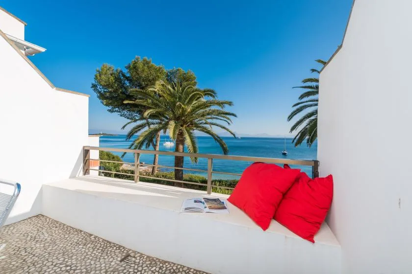 Una luminosa terraza junto al mar, con cojines rojos y palmeras frente al agua.